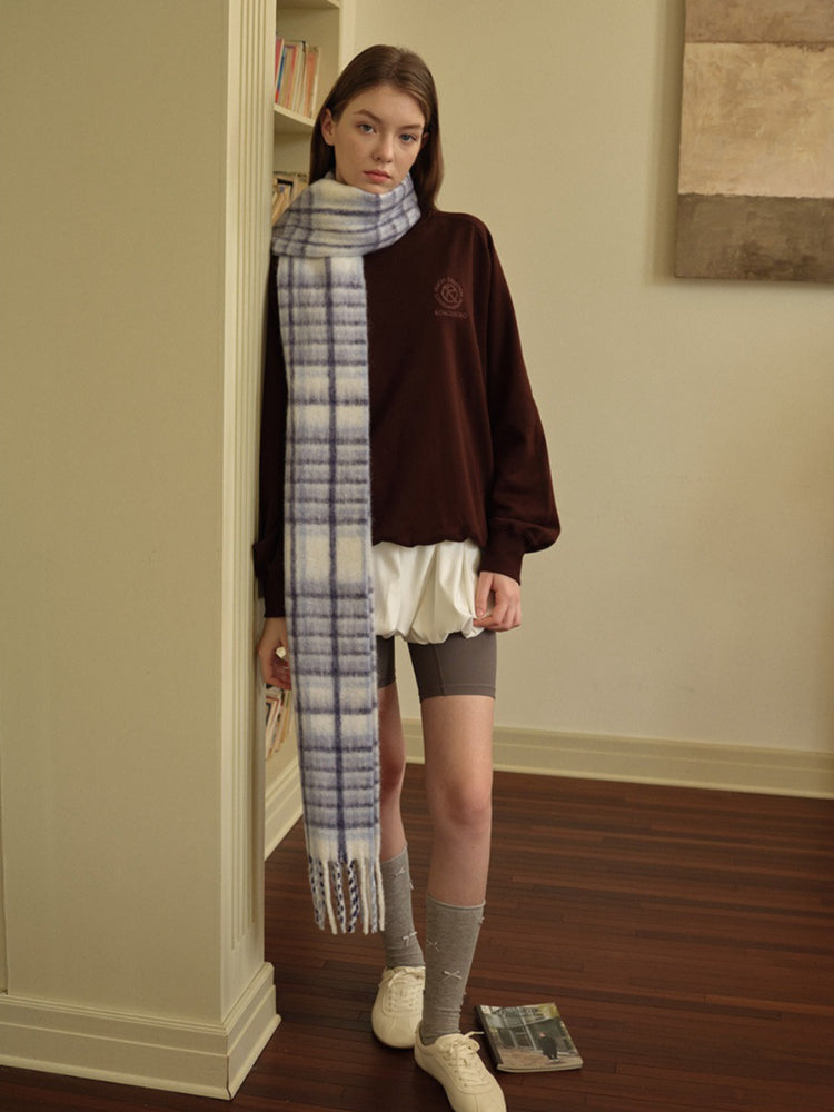 A young woman stands in front of a bookshelf wearing a brown sweatshirt, a white ruffled skirt, and a red plaid scarf with fringed ends.