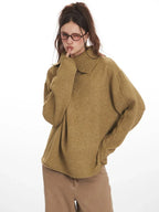 Close-up of a model wearing a tan oversized knit sweater with a wide collar and button detail, looking over her shoulder and winking.