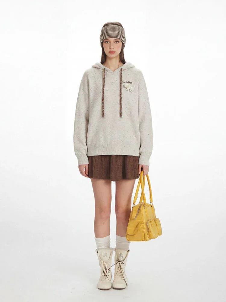 Model wearing a light-colored speckled knit hoodie with a brown pleated skirt, white socks, and cream-colored lace-up boots, holding a yellow handbag.