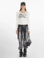 Woman wearing a white long-sleeved graphic t-shirt, grey distressed jeans with a black fringed belt, black boots, a black beaded necklace, and a black and white polka dot baseball cap, holding a black handbag.