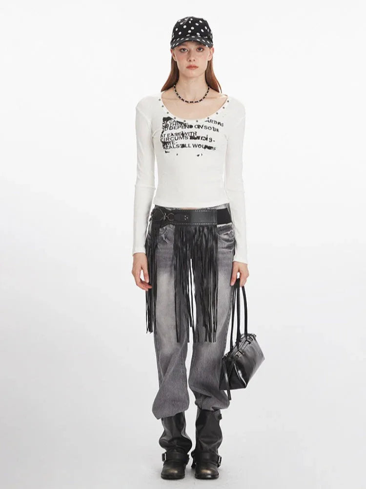 Woman wearing a white long-sleeved graphic t-shirt, grey distressed jeans with a black fringed belt, black boots, a black beaded necklace, and a black and white polka dot baseball cap, holding a black handbag.
