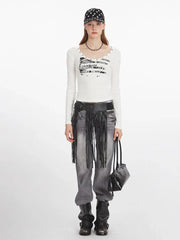 Woman wearing a white long-sleeved graphic t-shirt, grey distressed jeans with a black fringed belt, black boots, a black beaded necklace, and a black and white polka dot baseball cap, holding a black handbag.