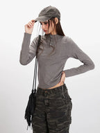 Close-up of a woman wearing a grey long-sleeved mock neck top with button detailing and a cropped hem, paired with camouflage print wide-leg cargo pants, and a grey baseball cap.