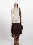 Model wearing a cream ribbed turtleneck sweater, brown asymmetrical hem skirt, cream belt, brown boots, cream knit beanie, and a brown faux fur shoulder bag, front view.