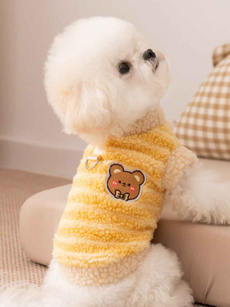 Side view of a white bichon frise dog wearing a yellow striped fleece vest with sherpa trim and a bear patch