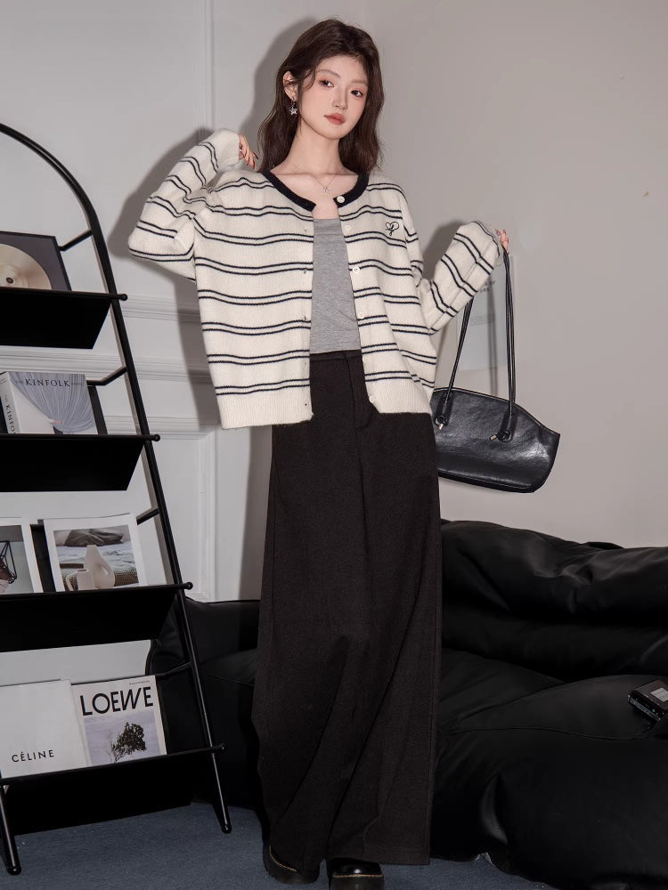 Full body shot of a woman wearing a cream and black striped button-up knit cardigan with dark wide-leg pants and holding a black handbag.