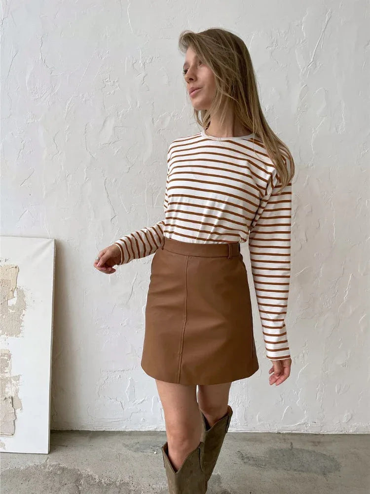 Woman wearing a white and brown striped long-sleeve top, a brown faux leather mini skirt, and brown boots.