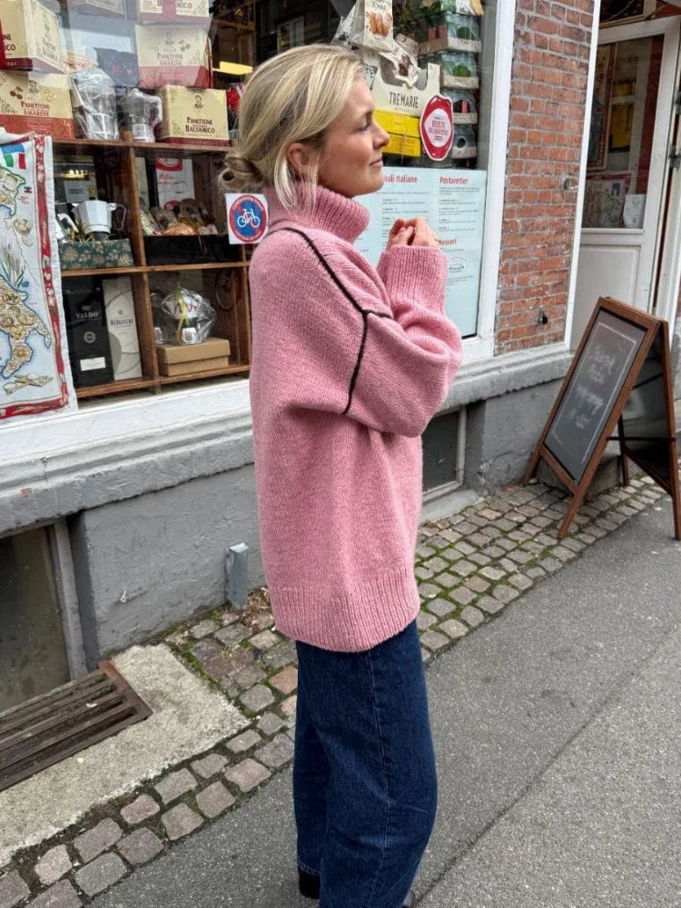 Woman wearing a pink oversized turtleneck sweater with contrasting black seam details and dark wash jeans, standing outside a shop. Main image