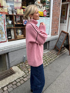 Woman wearing a pink oversized turtleneck sweater with contrasting black seam details and dark wash jeans, standing outside a shop.