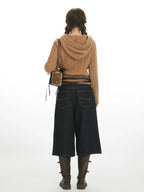 Back view of a woman wearing a brown cable knit hooded sweater with buttons and pom-poms, dark wash wide-leg cropped jeans, a black belt, and brown boots.