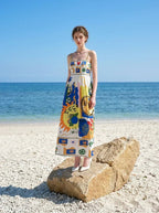 Woman wearing a white maxi dress with a colorful abstract print and spaghetti straps, standing on a rock on a sandy beach with the ocean in the background.