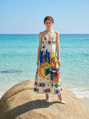 Woman wearing a white maxi dress with a colorful abstract print and spaghetti straps, standing on a large rock at the edge of the ocean.