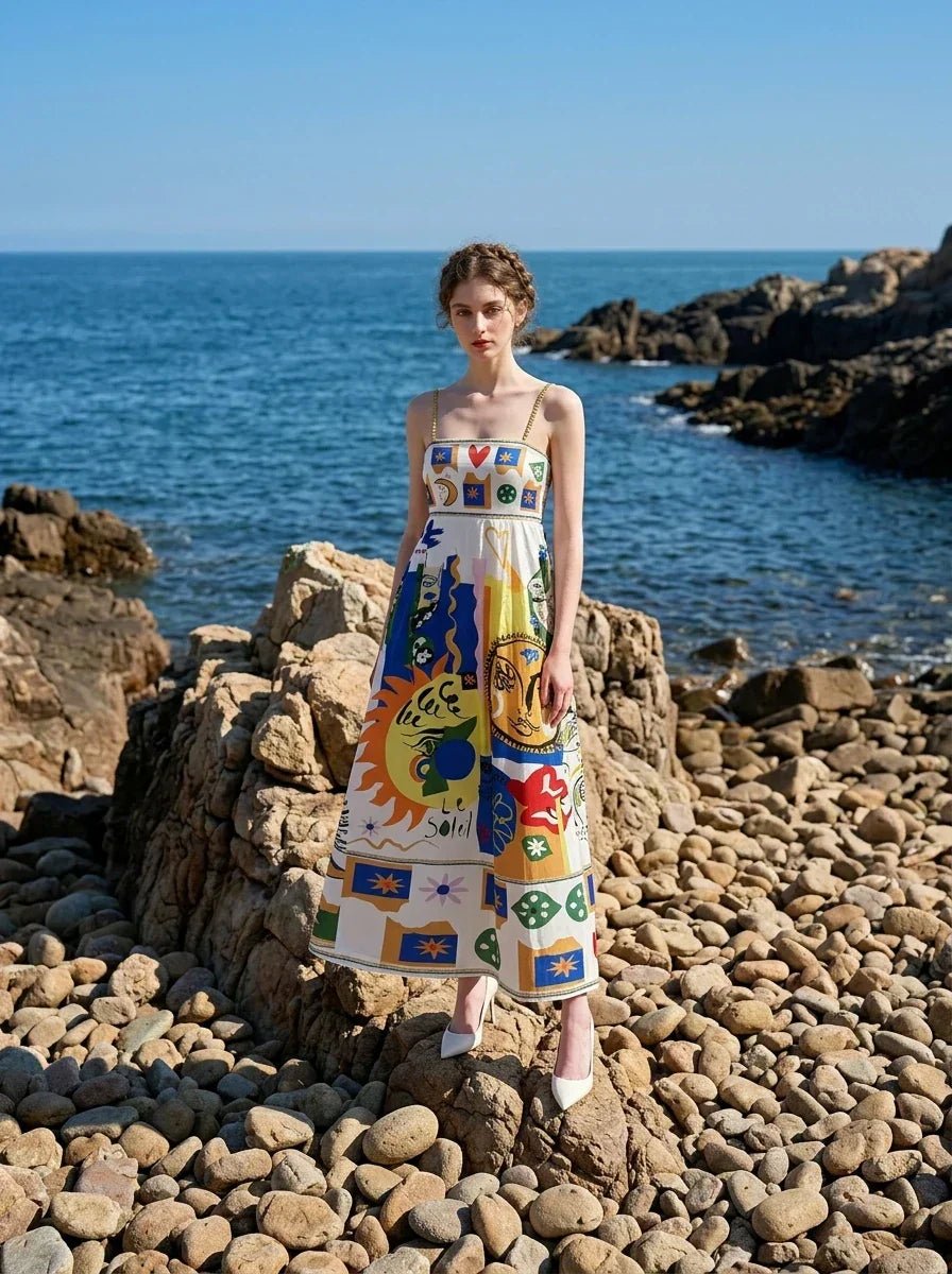 Woman wearing a white maxi dress with a colorful abstract print and spaghetti straps, standing on rocks by the sea.