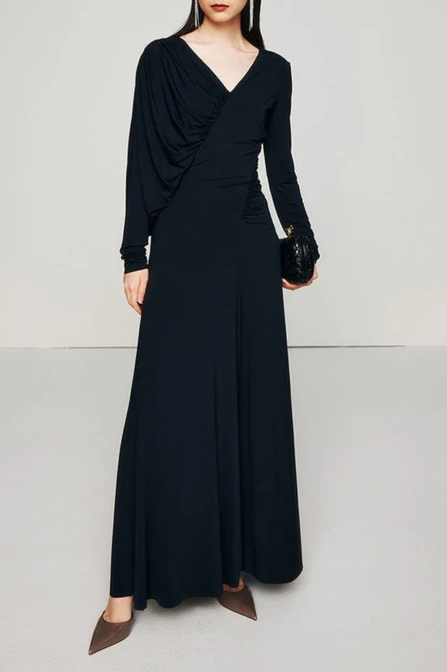 A woman models a long, black, long-sleeved dress with asymmetrical draping and ruching, holding a black clutch.