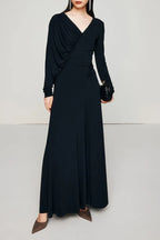 A woman models a long, black, long-sleeved dress with asymmetrical draping and ruching, holding a black clutch.