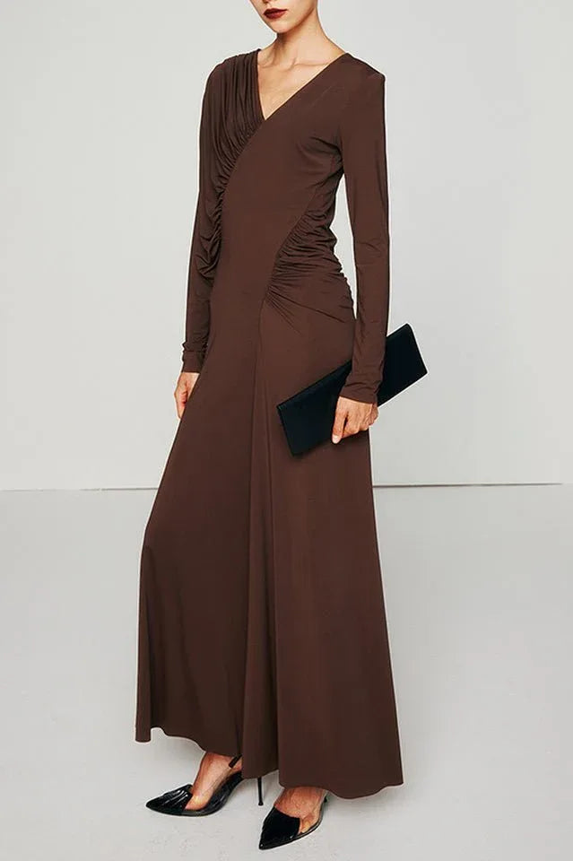 A woman models a long, brown, long-sleeved dress with asymmetrical draping and ruching, holding a black clutch.