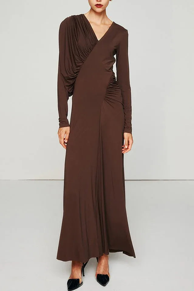 A woman models a long, brown, long-sleeved dress with asymmetrical draping on the left shoulder and ruching on the right side of the bodice.