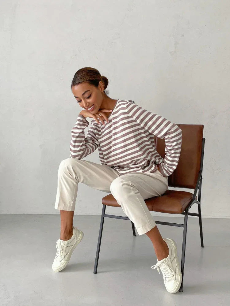 Woman sitting on a chair wearing a brown and white striped long-sleeved crew neck sweater with cream-colored pants and white sneakers, resting her chin on her hand.