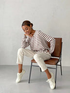 Woman sitting on a chair wearing a brown and white striped long-sleeved crew neck sweater with cream-colored pants and white sneakers, resting her chin on her hand.