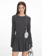 Woman wearing a dark grey long-sleeve mini dress with a gathered waist, white-trimmed grey socks, and silver shoes, smiling and holding a silver handbag and a small striped pouch around her neck.