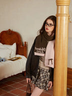 Model in a pink and green knit scarf with embroidered text, wearing a leather jacket and plaid skirt, leaning against a wooden pillar.