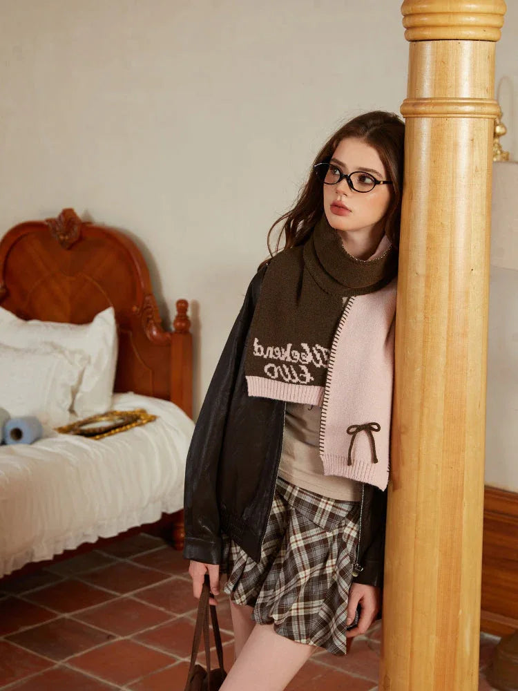 Model in a pink and green knit scarf with embroidered text, wearing a leather jacket and plaid skirt, leaning against a wooden pillar.