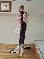 A young woman pulls her dark scarf closer to her face, covering her chin. She is dressed in a pink knit cardigan, white pleated skirt, white tights, leg warmers, and pink ballet flats.