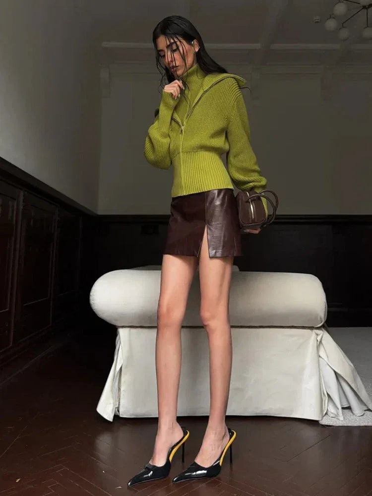 Model posing in a lime green zip-up knit sweater, brown leather mini skirt, and black heels, holding a brown handbag.