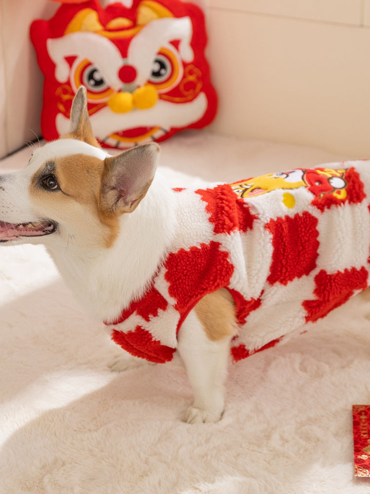 Side view of a corgi dog wearing a red and white checkered plush sweater, with a decorative pillow and lion head cushion in the background.