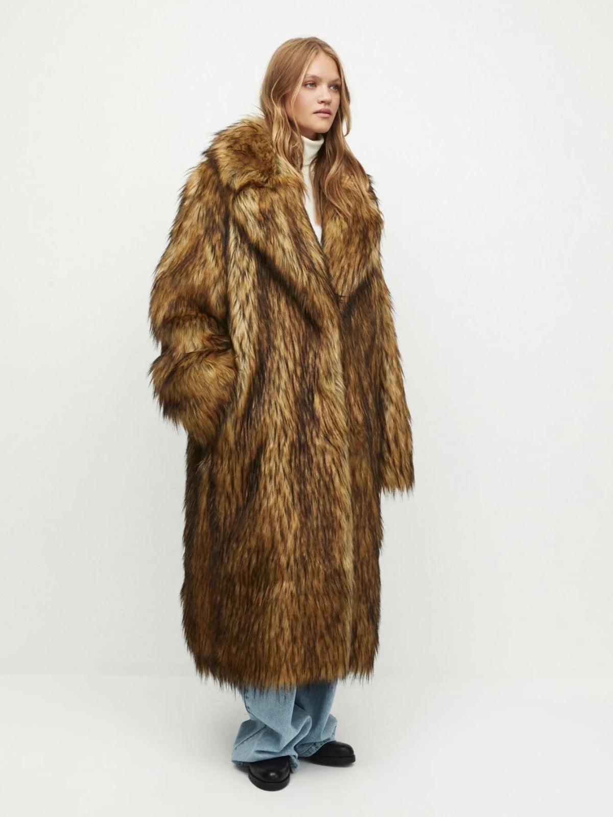 Woman wearing a long brown faux fur coat with a white turtleneck and blue jeans, side view.