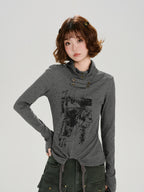 Woman wearing a grey long-sleeve graphic print turtleneck top with thumbhole cuffs and a drawstring hem, posing with one hand on her hip.