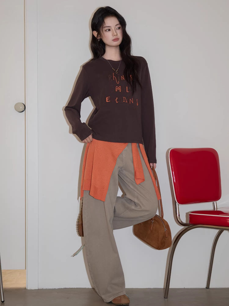 Full body shot of a young woman wearing a brown long-sleeved graphic t-shirt and beige pants, leaning against a red chair