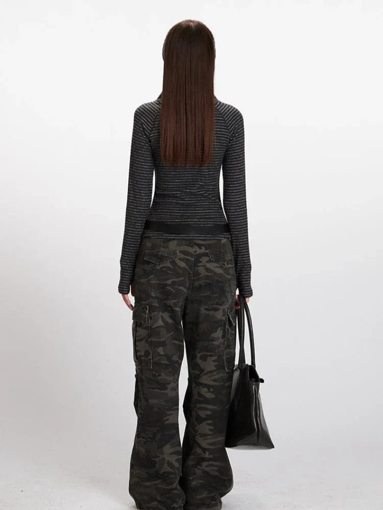 Back view of a model wearing a gray striped long-sleeve top with a mock neck and thumbholes, paired with camouflage cargo pants and a black belt, holding a black tote bag.