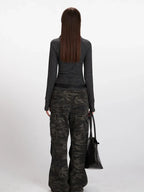 Back view of a model wearing a gray striped long-sleeve top with a mock neck and thumbholes, paired with camouflage cargo pants and a black belt, holding a black tote bag.