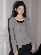 Woman wearing a grey long-sleeved top with a draped overlay and black pants, standing against a wall.