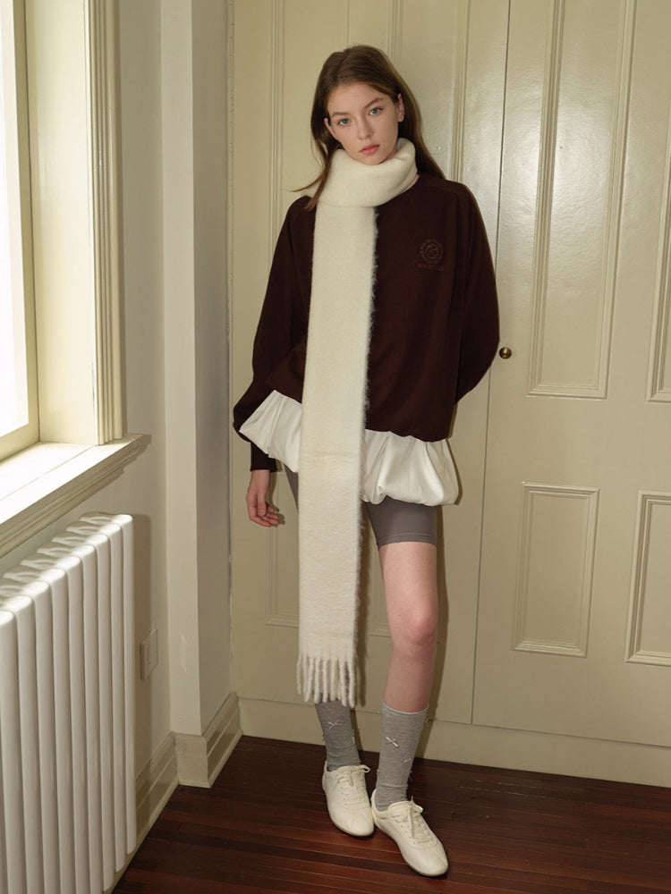 Woman standing in a doorway, wearing a brown velour sweatshirt with a white scarf, grey shorts, grey socks, and white sneakers.