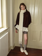 Woman standing in a doorway, wearing a brown velour sweatshirt with a white scarf, grey shorts, grey socks, and white sneakers.