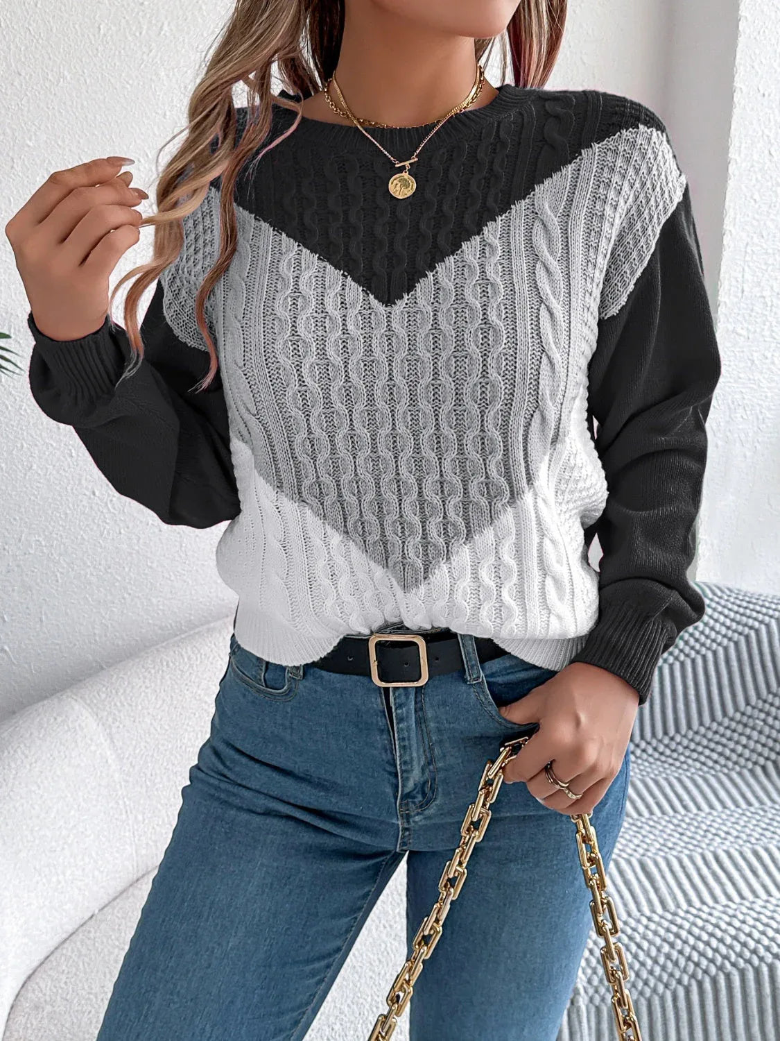 Woman wearing a black, grey, and white color block chevron cable knit sweater and blue denim jeans, accessorized with a gold chain necklace and a black belt.
