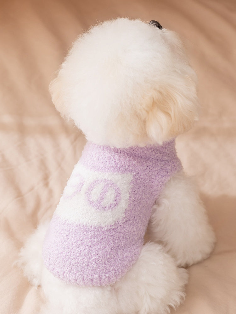 The back of a white bichon frise dog wearing a lavender plush dog sweater with a white graphic design, sitting on a beige surface.