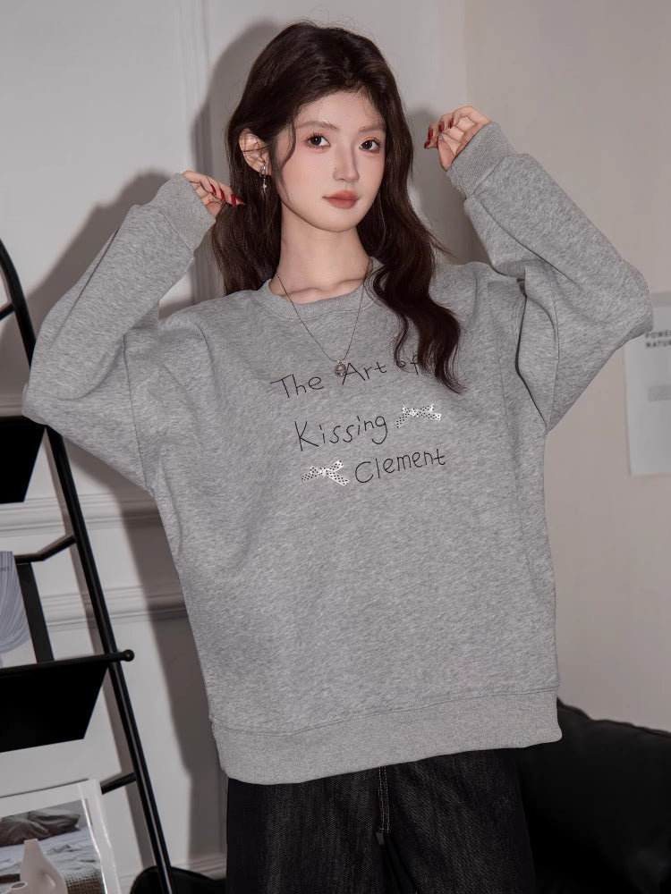 Woman in a grey sweatshirt with graphic print, posing with hands near ears