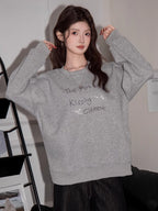 Woman in a grey sweatshirt with graphic print, posing with hands near ears