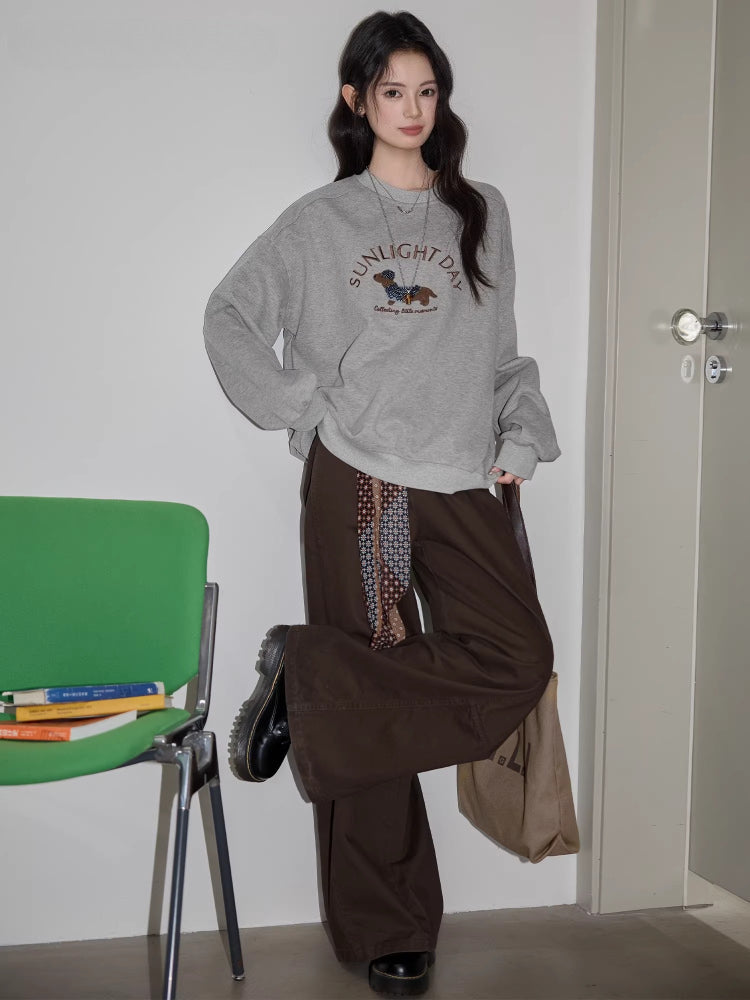 Full-body shot of a woman standing, wearing a grey crew neck sweatshirt with a dachshund graphic and text "SUNLIGHT DAY", brown wide-leg pants, and holding a tote bag.