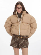 Front view of a model wearing a tan cropped puffer jacket, brown plaid pleated mini skirt, white leg warmers, and brown loafers.