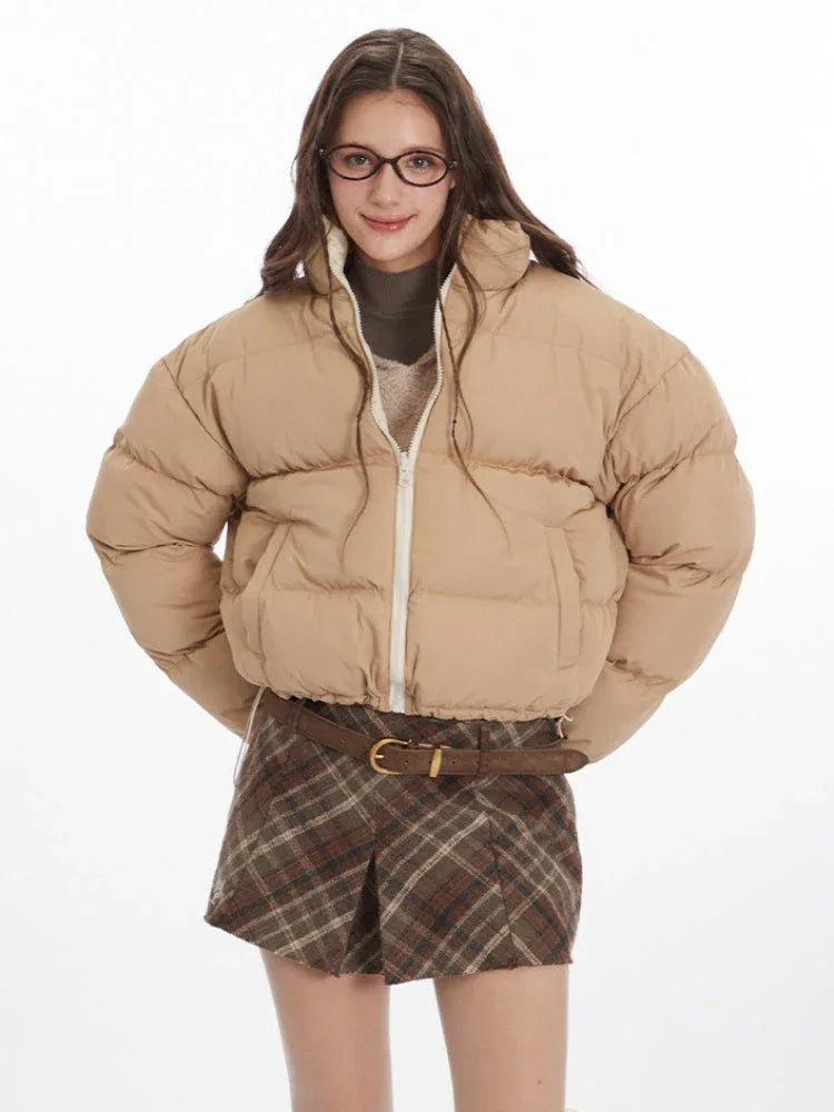 Front view of a model wearing a tan cropped puffer jacket, brown plaid pleated mini skirt, white leg warmers, and brown loafers.