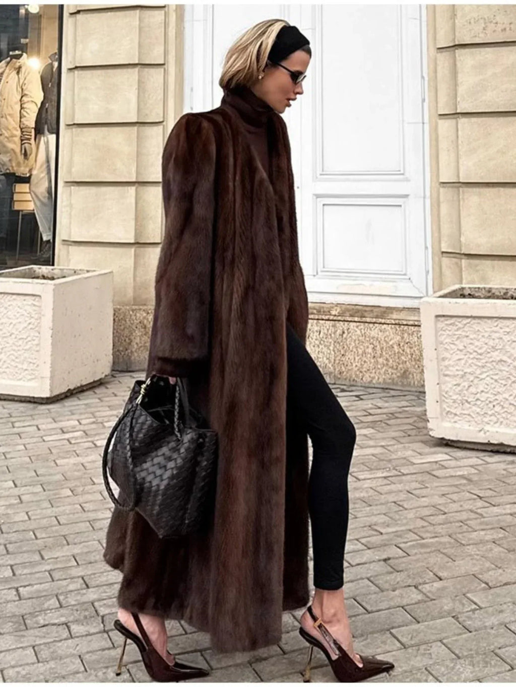 Woman wearing a long, brown mink fur coat, black turtleneck, black leggings, black woven handbag, and brown pointed-toe heels, walking on a cobblestone street.