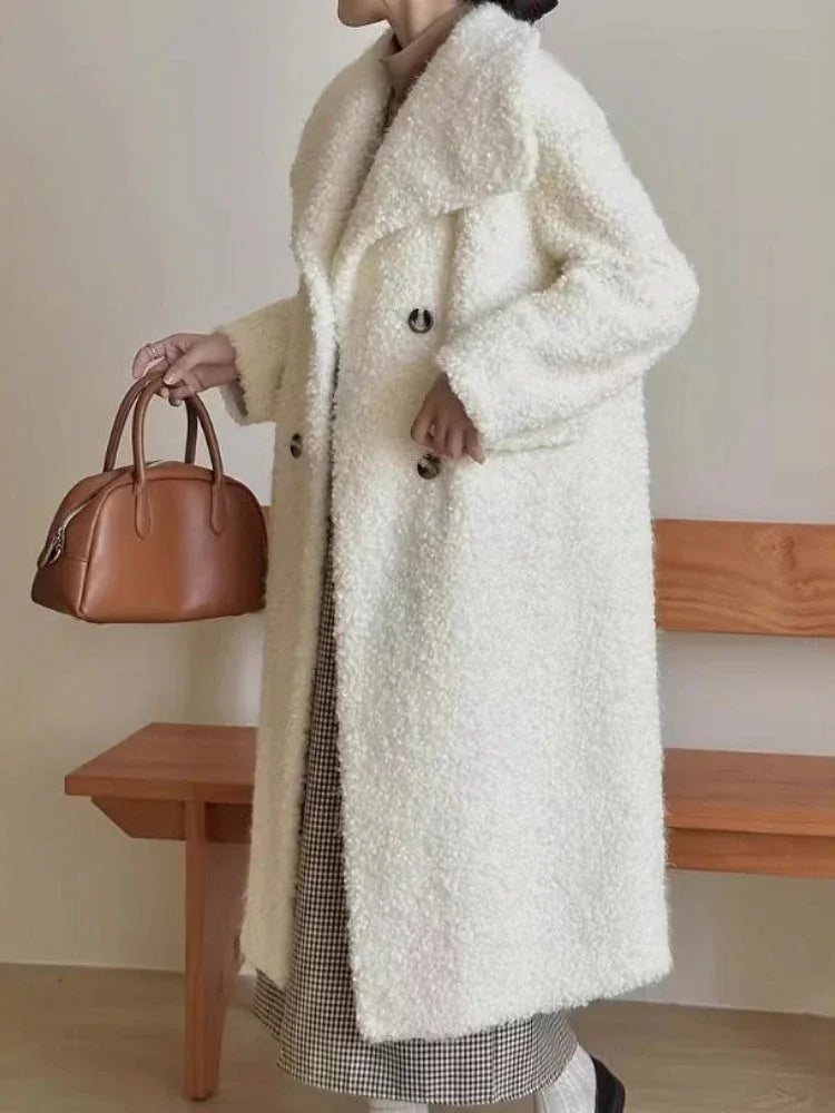 Woman wearing a long white fuzzy fleece coat with a notched lapel and double-breasted closure, holding a brown handbag, standing next to a wooden bench.