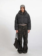 Model wearing a cropped grey puffer jacket with a hood, grey long-sleeved shirt, and camouflage cargo pants, holding a black tote bag.