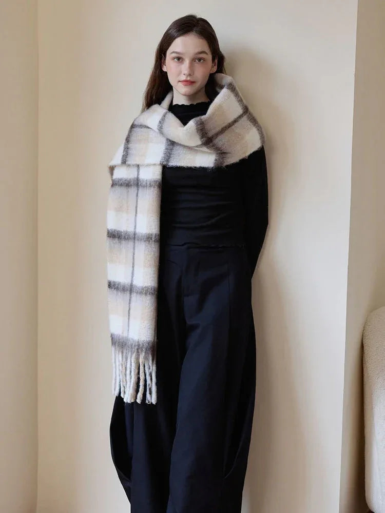 Full-body shot of a woman standing near a wall, wearing a black top and pants, with a prominent plaid scarf draped around her neck.