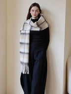Full-body shot of a woman standing near a wall, wearing a black top and pants, with a prominent plaid scarf draped around her neck.