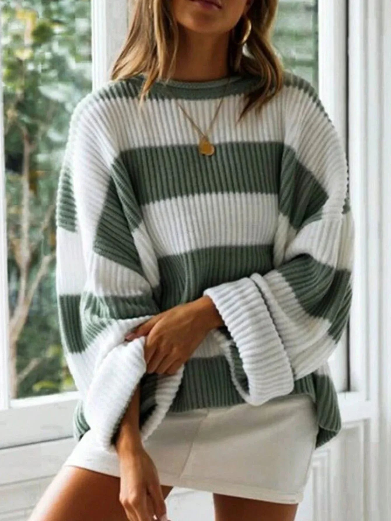 Woman wearing a green and white striped knit sweater with a crew neck and rolled-up sleeves, standing in front of a window, with one hand tucked into her sweater.